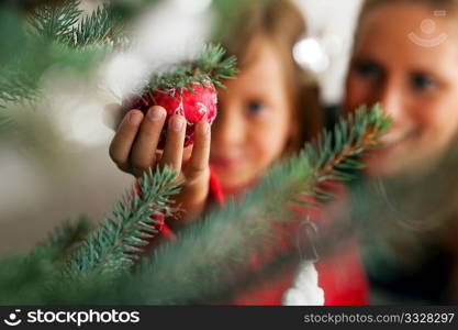Young girl helping her mother decorating the Christmas tree, holding some Christmas baubles in her hand (Focus on bauble)