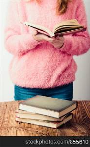 Young girl holding a book standing in bookstore. A few books on a wooden table. Teenager girl wearing pink sweater and blue jeans. Vertical photo
