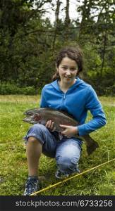 Young girl holding trophy rainbow trout with trees and sky in background