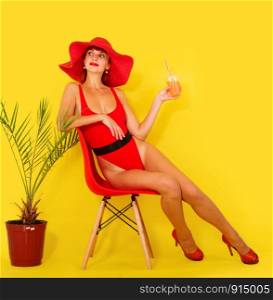 young girl in a bright red swimsuit and a wide-brimmed red hat drinks a cocktail while sitting on a chair on a yellow background. girl drink coktail