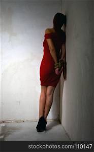young girl in a red dress with her hands tied behind her back is standing in the corner of a dilapidated shabby room