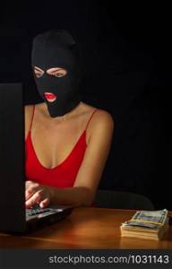 young girl in a sexy red dress and a balaclava mask sits at a laptop and makes an honest profit on the Internet. internet burglar in mask