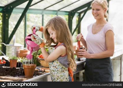Young girl in greenhouse watering plant with woman holding pot smiling
