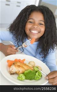 Young girl in kitchen eating chicken and vegetables smiling
