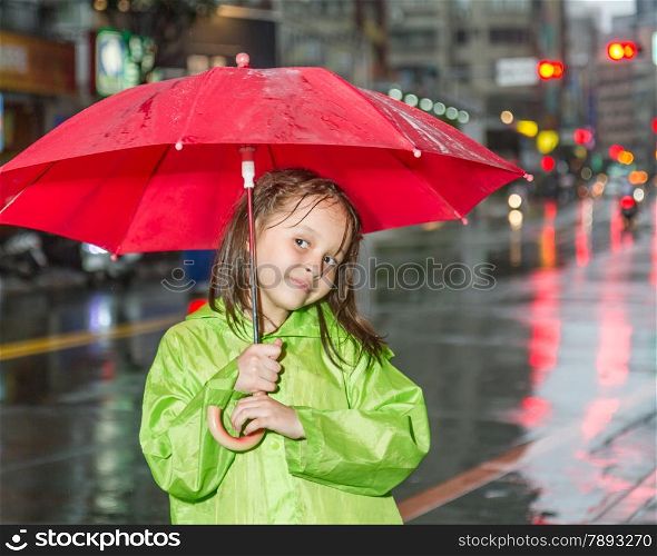 Young girl in rain wearing a green raincoat and holding a red umbrella by city street