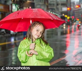 Young girl in rain wearing a green raincoat and holding a red umbrella by city street