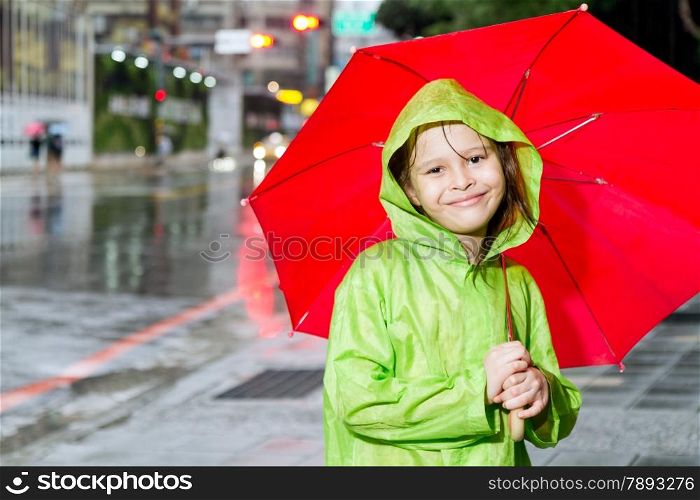 Young girl in rain wearing a green raincoat and holding a red umbrella on sidewalk next to street