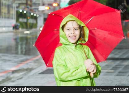 Young girl in rain wearing a green raincoat and holding a red umbrella on sidewalk next to street