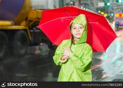 Young girl in rain wearing a green raincoat and holding a red umbrella, on sidewalk