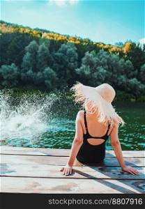 Young girl in straw hat and black swimsuit sitting on wooden bay near river at summer. Travel, relax, vacation concept,