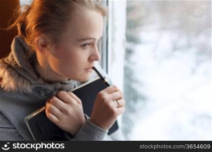 Young girl is dreaming by the window with notebook and pen