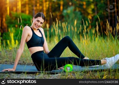 Young girl is exercising with a roller to relax muscles while sitting on the mat in the park. Young girl exercising with a roller to relax muscles