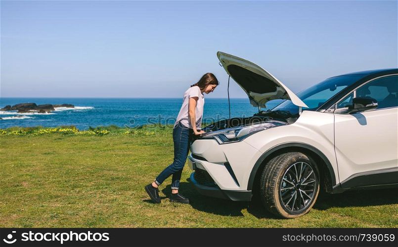 Young girl looking at the engine of her broken down car outdoors. Girl looking engine of broken down car