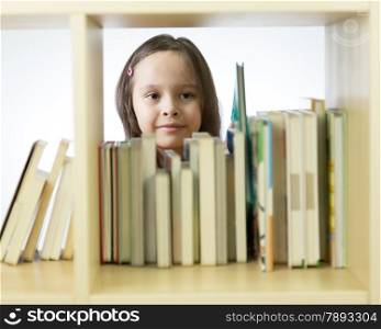 Young girl looking through books in library behind shelf. Horizontal