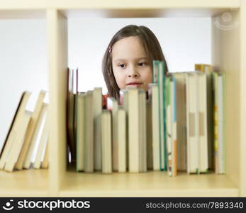 Young girl looking through books in library behind shelf. Horizontal