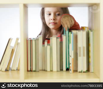 Young girl looking through books in library behind shelf. Horizontal