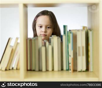 Young girl looking through books in library behind shelf. Horizontal
