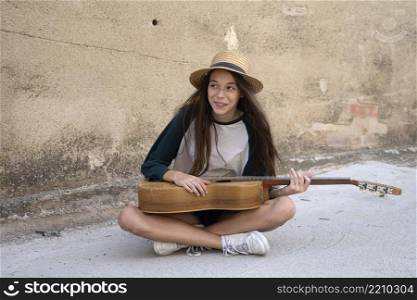 Young girl musician in summer clothes sitting on city ground playing acoustic guitar.