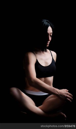 young girl of athletic appearance with black hair sits in a lotus position in a black bra and shorts, lateral twist exercise, dark background