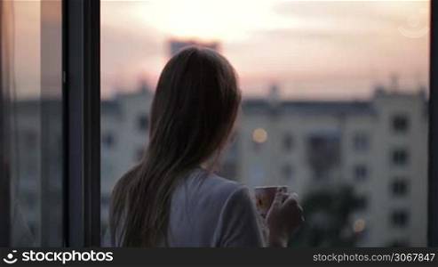 Young girl on the balcony watching the sunset and drinking tea. Middle shot