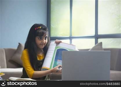 Young girl pointing towards a book in her hand while sitting in front of a laptop during online class