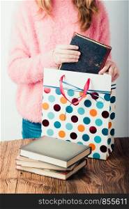 Young girl putting the books into paper bag in bookstore. A few books on a wooden table. Teenager girl wearing pink sweater and blue jeans. Vertical photo