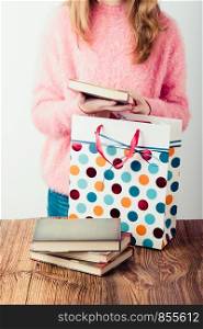 Young girl putting the books into paper bag in bookstore. A few books on a wooden table. Teenager girl wearing pink sweater and blue jeans. Vertical photo