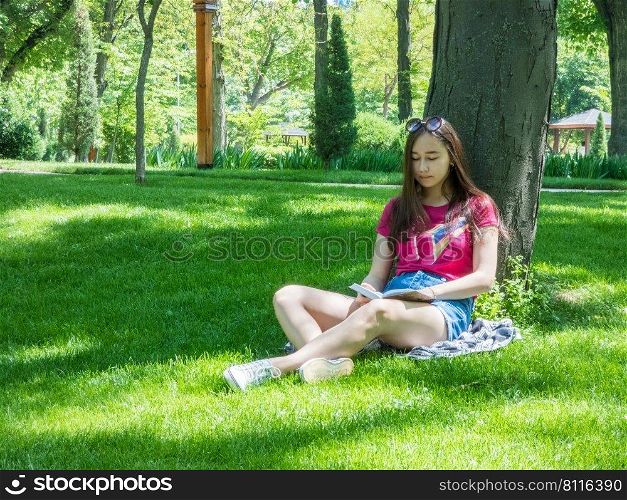 young girl reading a book in a picturesque park, sunny day. the girl of Asian appearance. young girl in a picturesque park