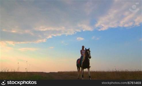 Young girl riding horse across the field in evening wide angle
