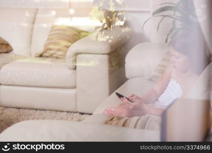 Young girl sitting in a sofa and using phone in living room behind the glass window