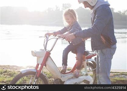 Young girl sitting on father&acute;s moped, father standing beside her, beside lake