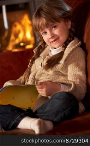Young Girl Sitting On Sofa And Reading Book By Cosy Log Fire