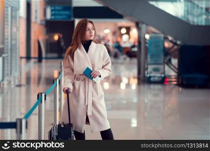 Young girl standing with a suitcase, holding a passport in the background of escalator at the airport.