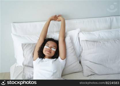 Young girl stretching in bed after wake up with moisturizing under eye patches on face. Pretty lazy teen lady lying in bedroom, enjoying morning skincare routine. Skin care, beauty, healthy habits.. Young girl stretching in bed after wake up with under eye patches on face. Skin care, healthy habits