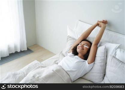 Young girl stretching lying in bed with cosmetic under eye patches on face skin. Happy biracial teen lady stretch after awaking in bedroom, enjoying skincare, morning beauty routine.. Girl stretching lying in bed with under eye patches on face skin. Skincare, morning beauty routine