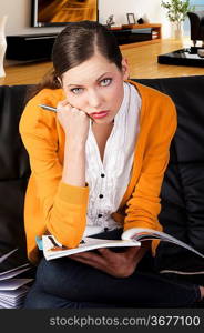 young girl student sitting on a sofa and reading a book, she is on her knees on the sofa, looks on to the lens with serious expression, her face is resting on right fist.