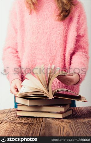 Young girl turning the pages of book in library A few books on a wooden table. Teenager girl wearing pink sweater and blue jeans. Vertical photo