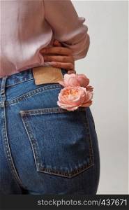 Young girl wearing of jean pants and pink shirt from back with natural flowers roses in a pocket on a gray background, copy space. Concept of Woman's or Mother's Day.. Perfect woman's bottom in a blue jeans and fresh roses living coral color in a back pocket on a light gray background, place for text. Congratulation card.