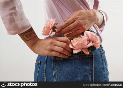Young girl wearing of jeans pants and pink shirt from back put in natural flowers roses in a pocket on a gray background, place for text. Concept of Woman's or Mother's Day.. Delicate girl's hands with tattoo put in the back pocket of blue jeans nice fresh roses coral living color on a light gray background, copy space.