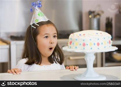 Young girl wearing party hat at kitchen counter looking at cake smiling