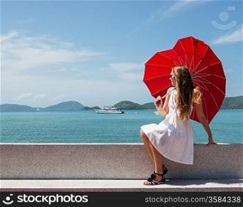 young girl with a red umbrella sits on the waterfront and looks at the sea