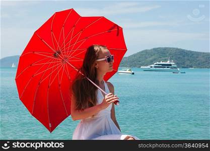 young girl with a red umbrella sits on the waterfront and looks at the sea