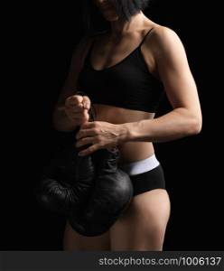 young girl with black hairs and a muscular body holds a pair of old black boxing gloves, she is dressed in a black bra and shorts, low key