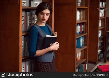 Young girl with book in hands standing between bookshelves at the school library.