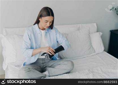 Young girl with disability setting her bionic prosthesis of arm sitting on bed at home. Disabled woman touching modern sensory artificial robotic hand in bedroom. Medicine technologies concept.. Modern young girl with disability setting her bionic prosthesis of arm sitting on bed at home