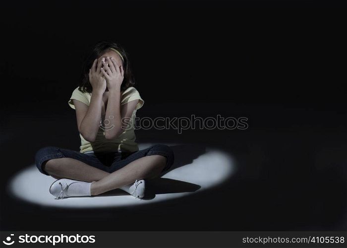 Young girl with hands covering her face sitting in spot light with dark background