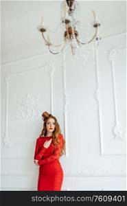 young girl with red hair in a bright red dress in a light room near a white wall