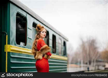 young girl with red hair in a bright red dress near an old green passenger car