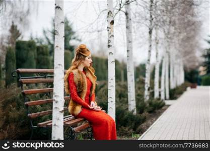 young girl with red hair in a bright red dress on a bench in an empty park among the birches