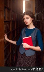 Young girl with the book in hands standing between book shelves
